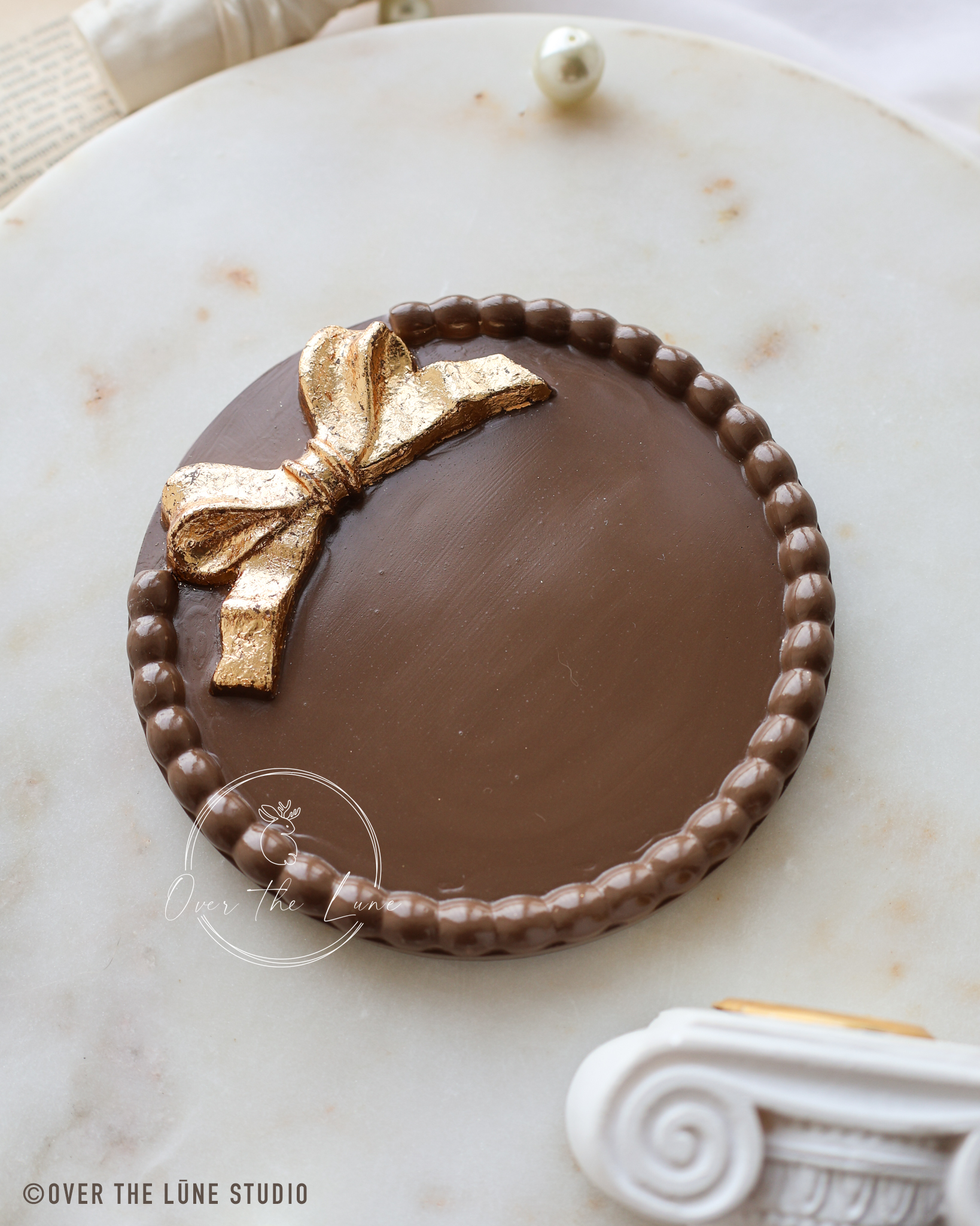 A decorative trinket tray sign with a golden bow design and a beaded border, displayed on a white marble surface. metallic gold bow decoration on top, photographed on a light marble surface.
