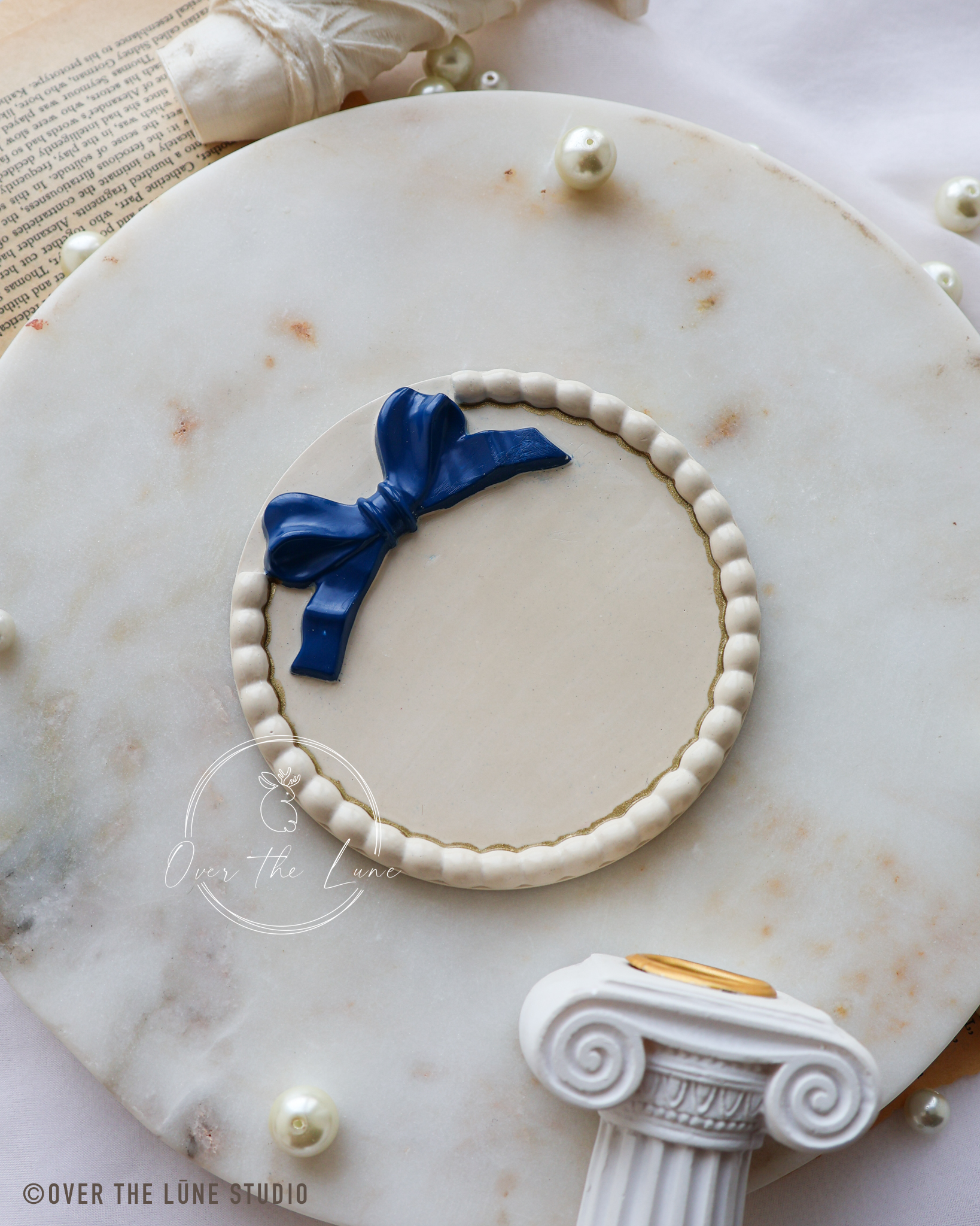 A circular white beaded wreath decorative tray with a dark blue ribbon bow, displayed on a white marble serving platter with decorative pearls and classical column props.