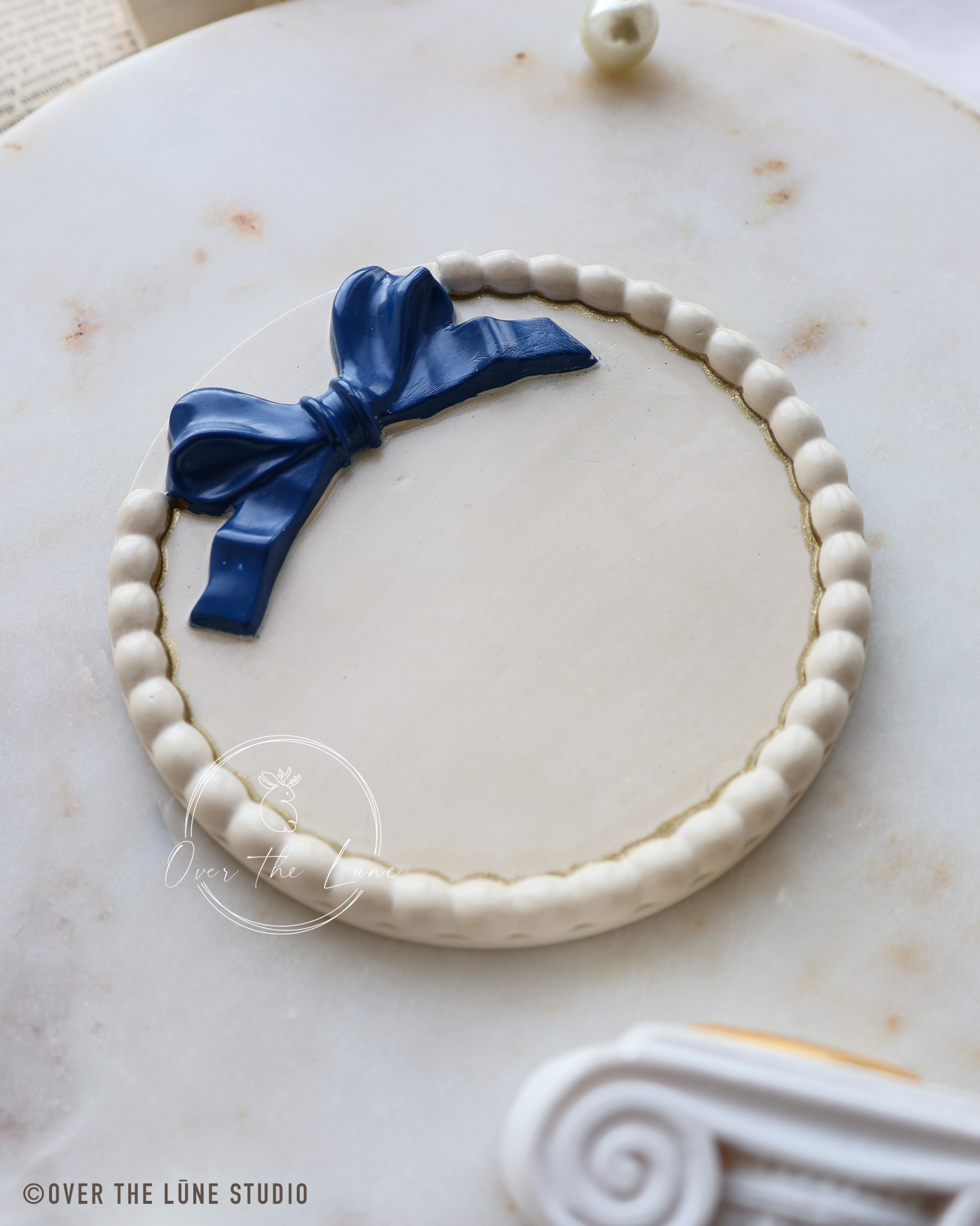 A round white plaque or sign with a beaded border and a glossy dark blue bow decoration, photographed on a light marble surface.