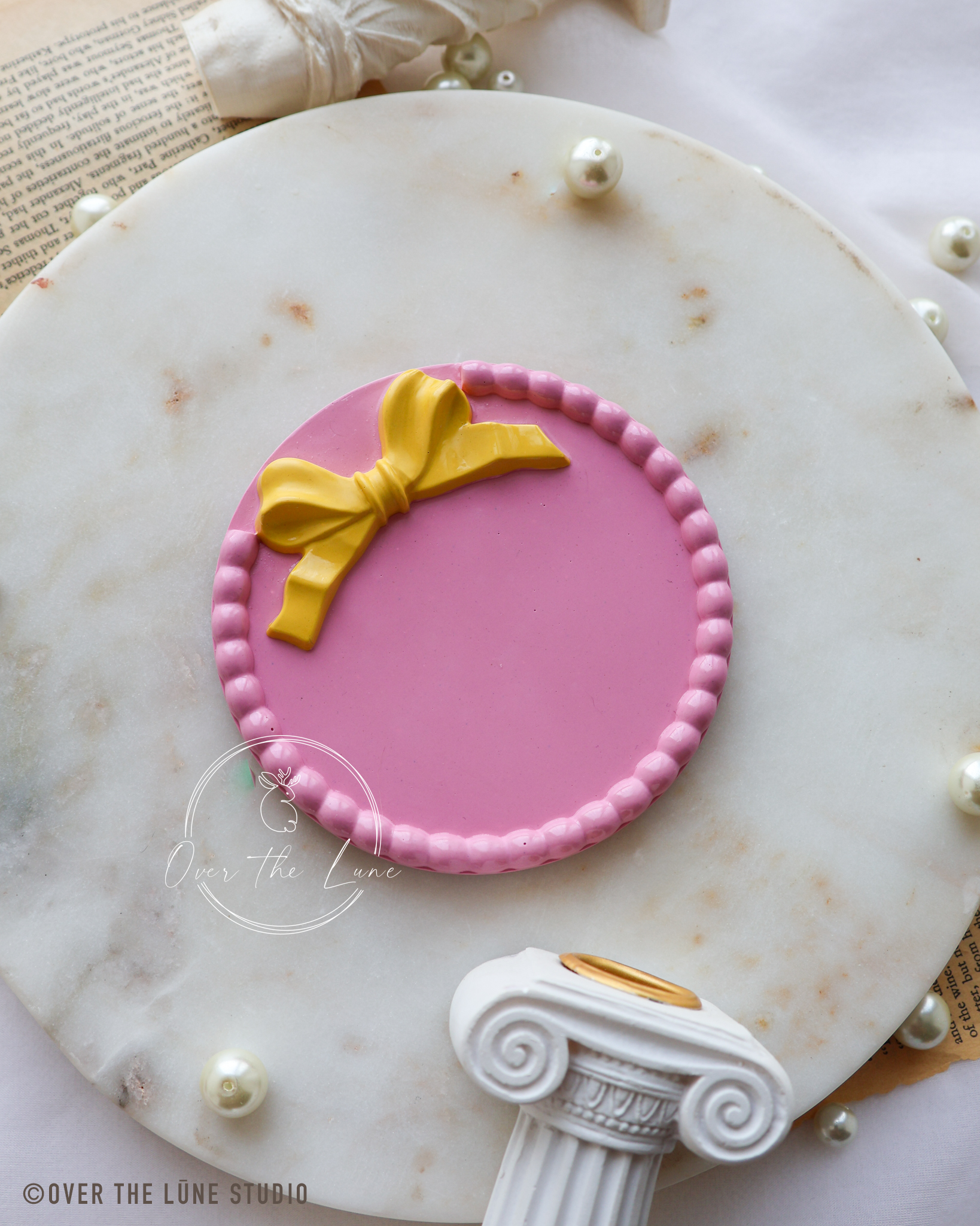 A pink round trinket tray with a yellow bow decoration, displayed on a white marble charger plate with pearl-like beads and decorative props.