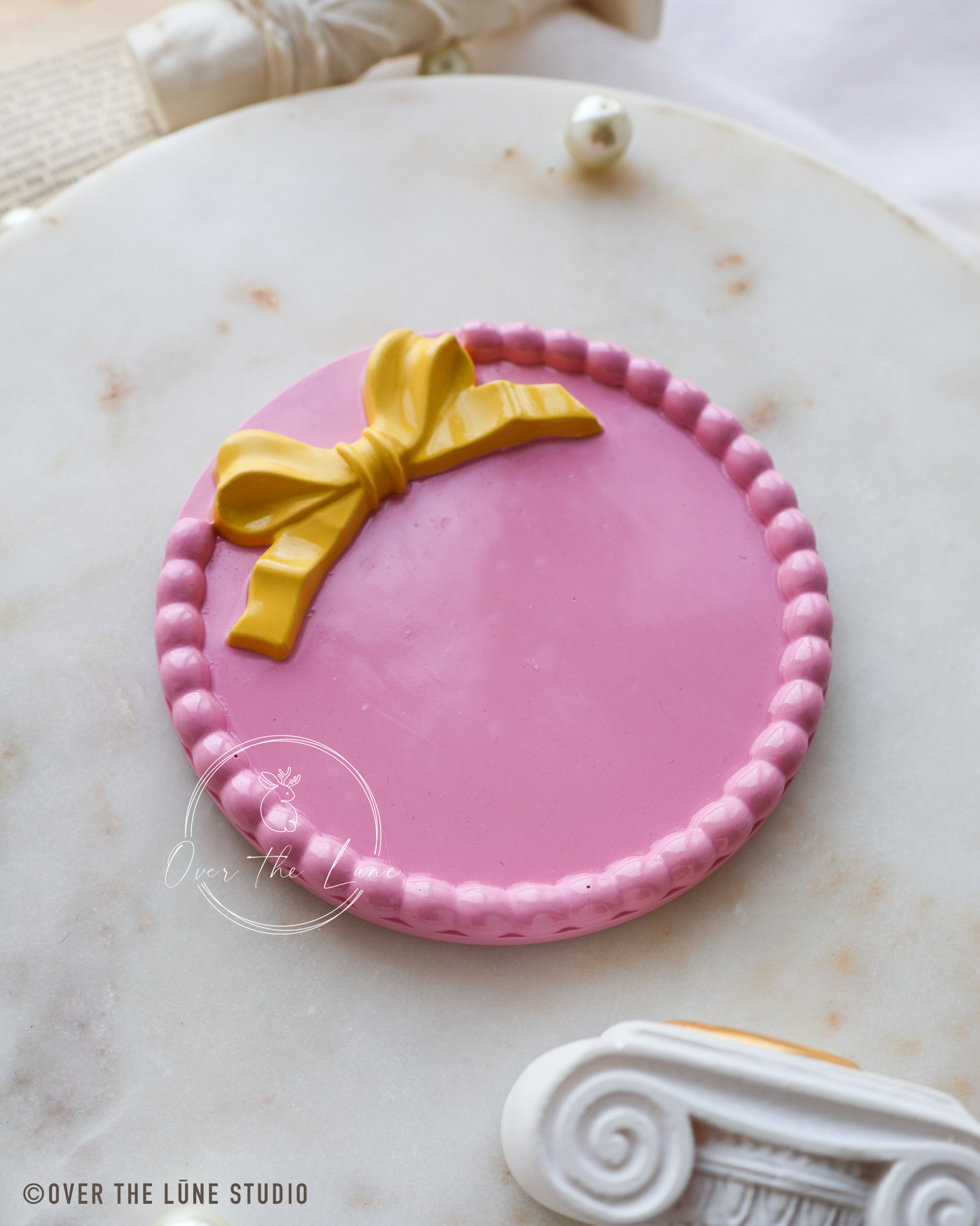A round pink decorative plaque or sign with a yellow bow design and a beaded border, displayed on a white marble surface.