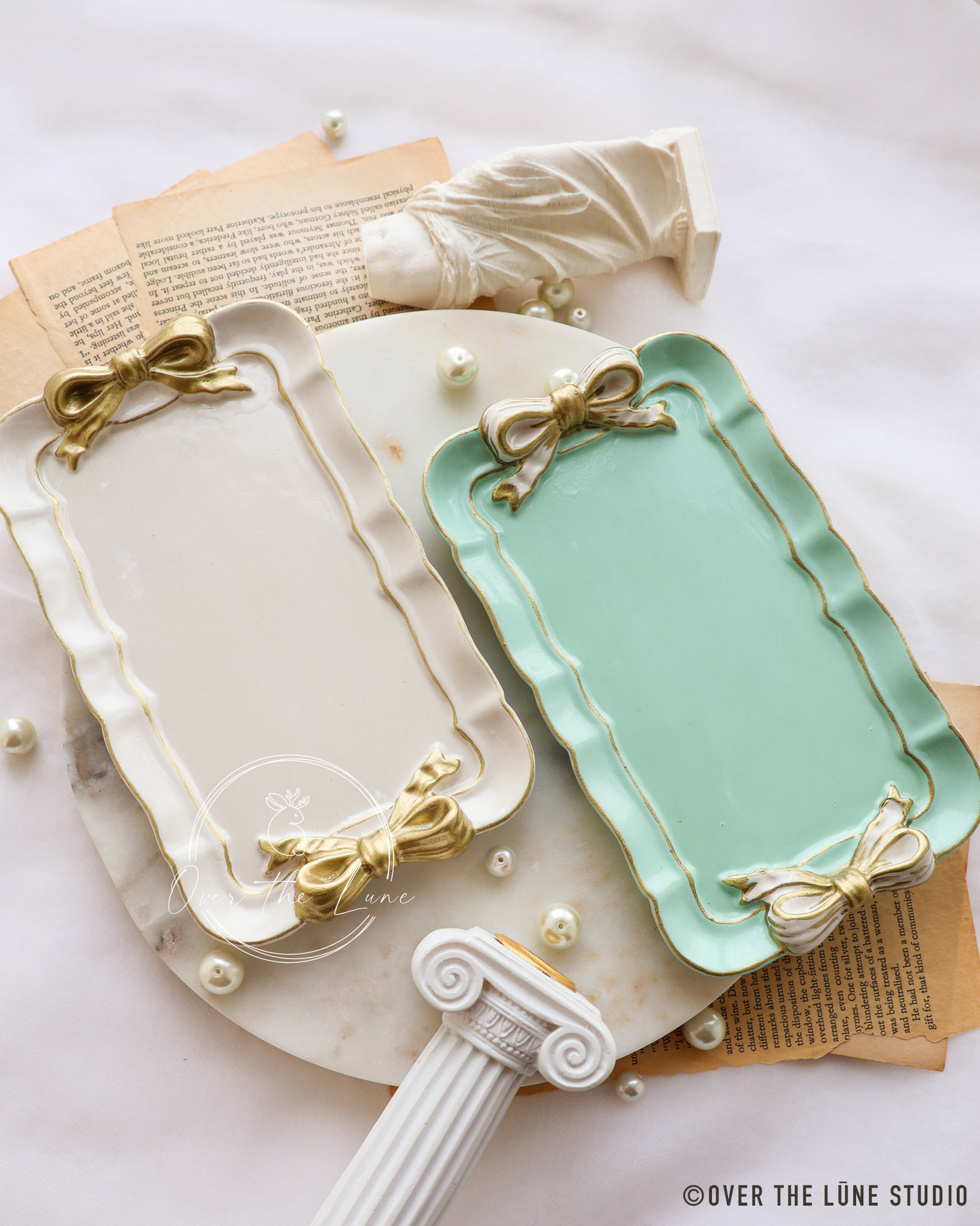 Two decorative rectangular trays with scalloped edges and bow details, one in warm cream and one in mint green, styled on a marble surface with vintage book pages, pearls, and a classical column prop.