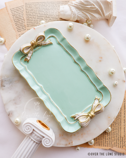 A mint green rectangular trinket tray with scalloped edges, gold trim, and decorative bow details on the corners, styled on a white marble charger plate with vintage book pages, pearls, and a white classical column prop.