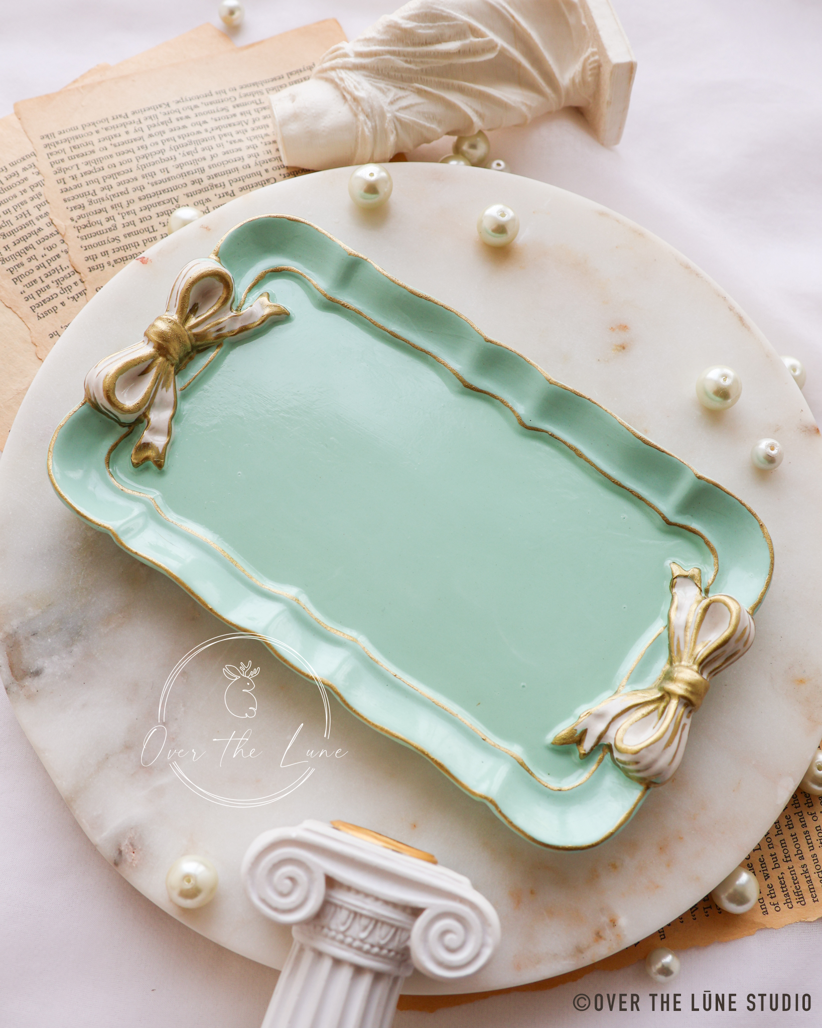 A mint green rectangular trinket tray with gold trim and decorative cream ribbon-bow details at the corners, styled on a round white marble platter with pearl-like beads and vintage book pages.