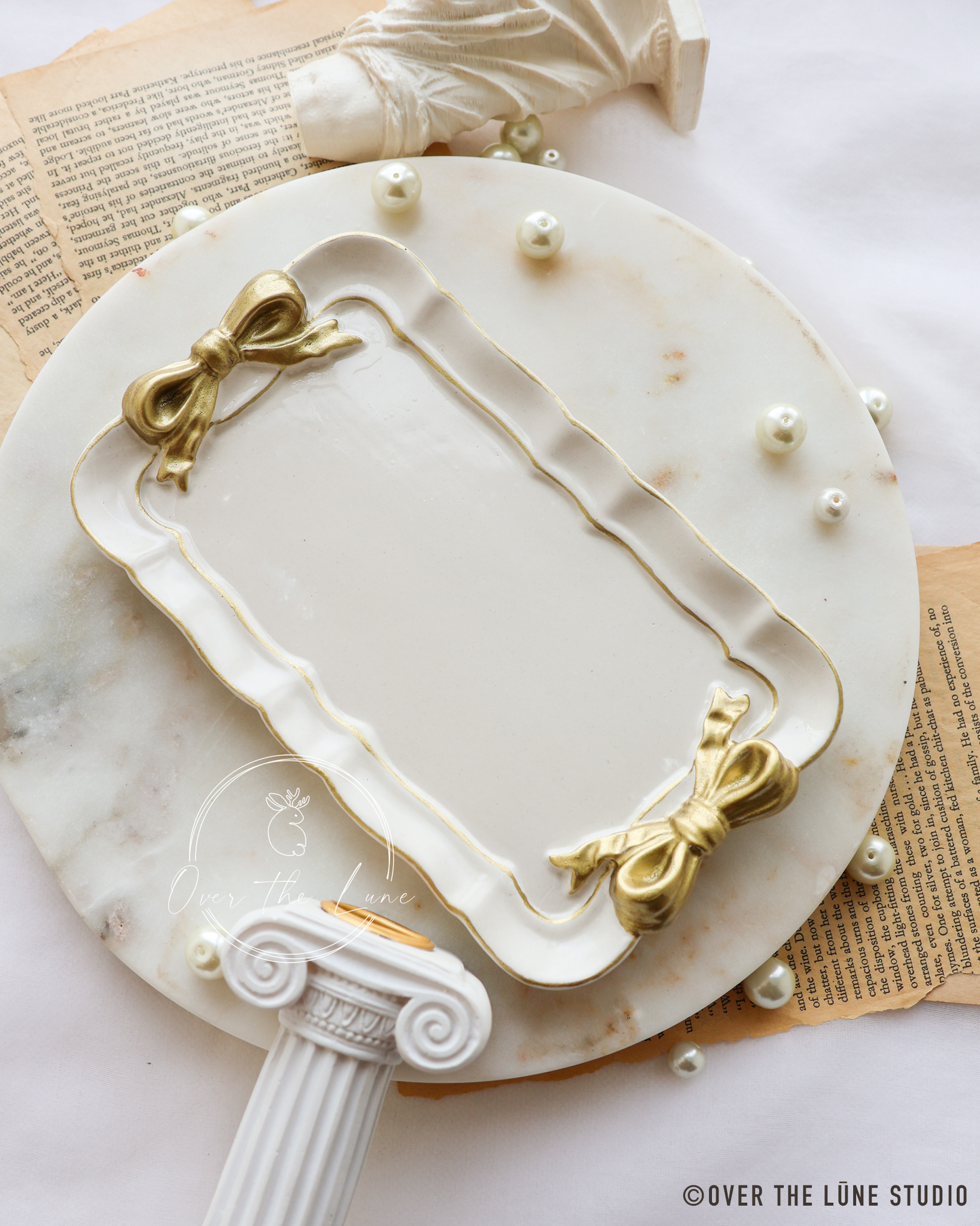 A decorative rectangular white tray with a gold scalloped border and gold bow-like corner accents, placed on a round white marble platter with pearl-like beads. The scene includes classical-style props, vintage paper, and a watermark reading OVER THE LUNE STUDIO.