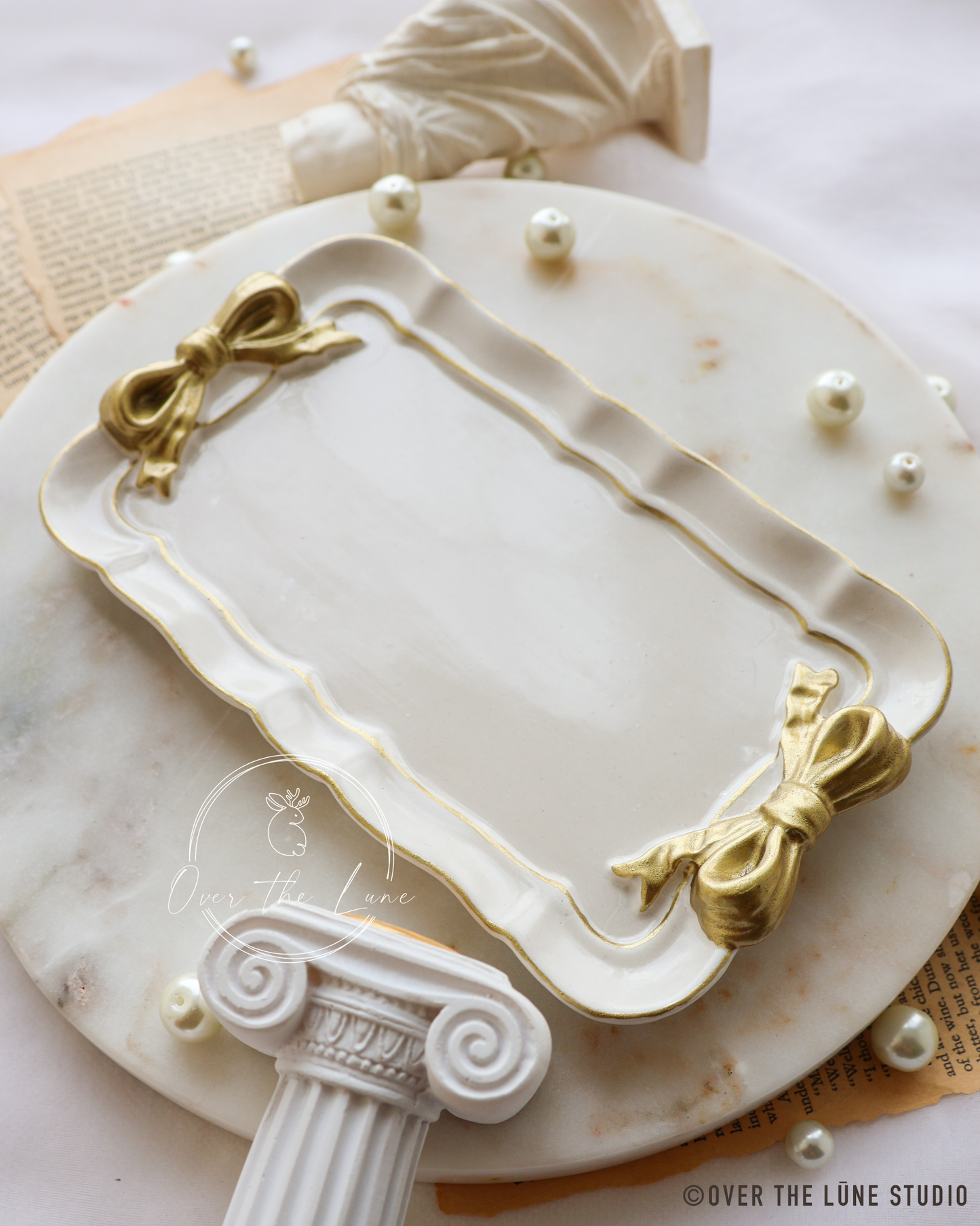 An ornate rectangular trinket tray with scalloped edges, gold bow details at the corners, and a white marble base, styled on a round marble platter with decorative props.