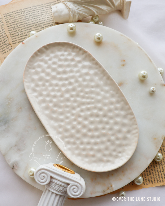 A white oval tray with a textured, dimpled surface, presented on a round white marble platter with decorative pearl-like beads and classical column accents.
