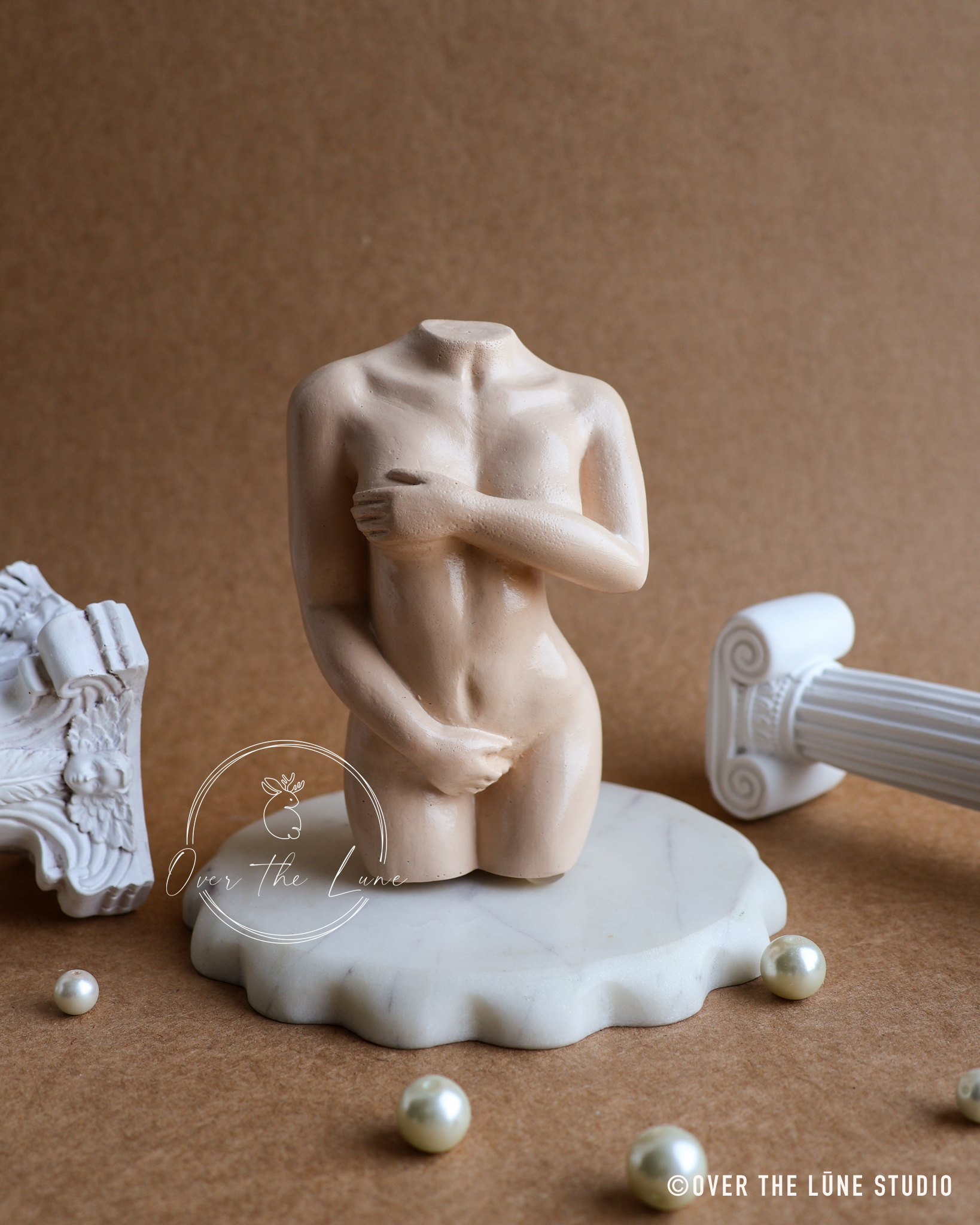 A small beige figurative sculpture of a headless nude female torso standing on a white marble base, styled on a brown background with decorative white classical fragments and pearl-like beads.