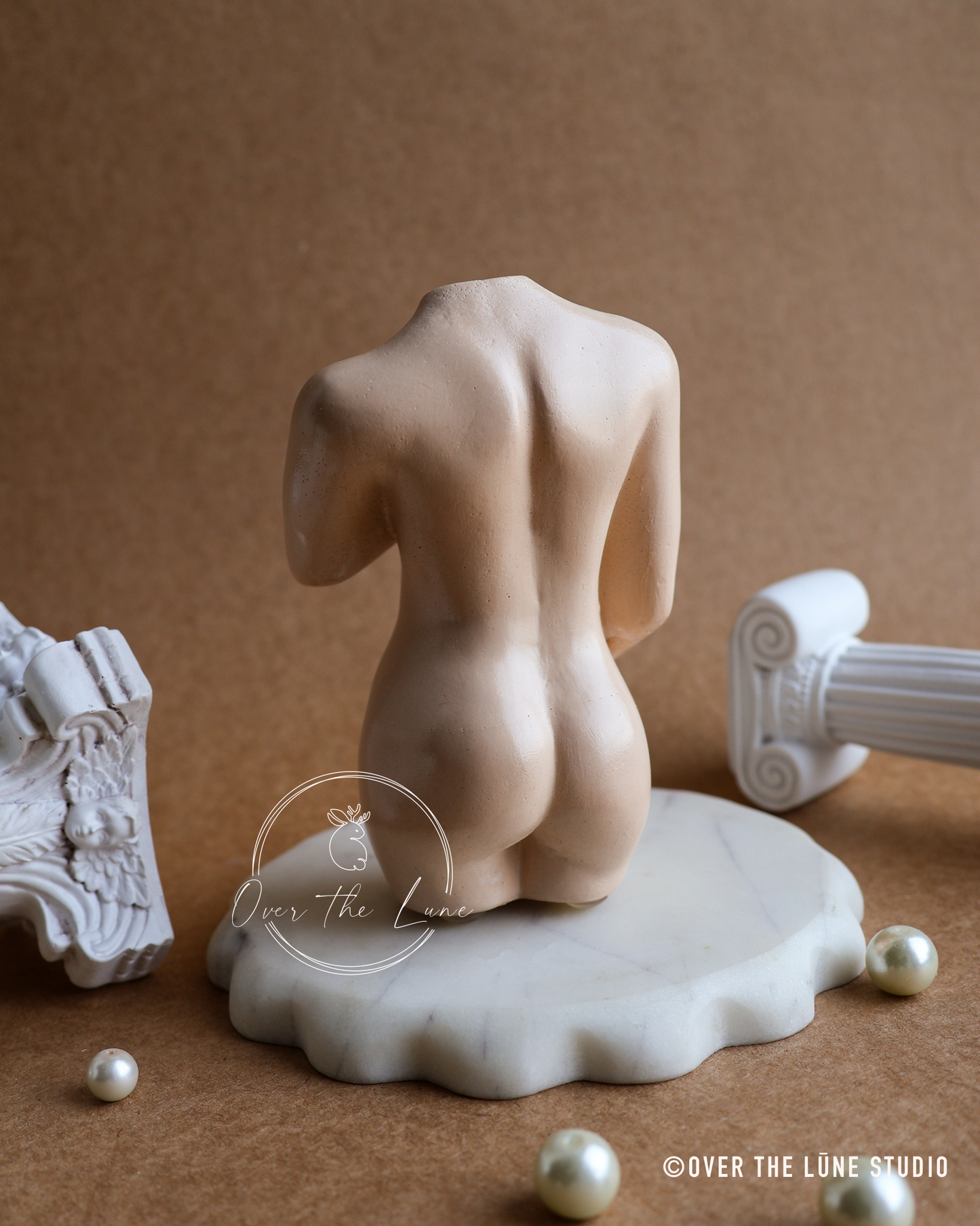 A beige plaster sculpture of a kneeling nude female figure on a white marble base, styled with classical column fragments and pearls on a brown background. Watermark 'Over the Lune' and '© OVER THE LUNE STUDIO' visible.