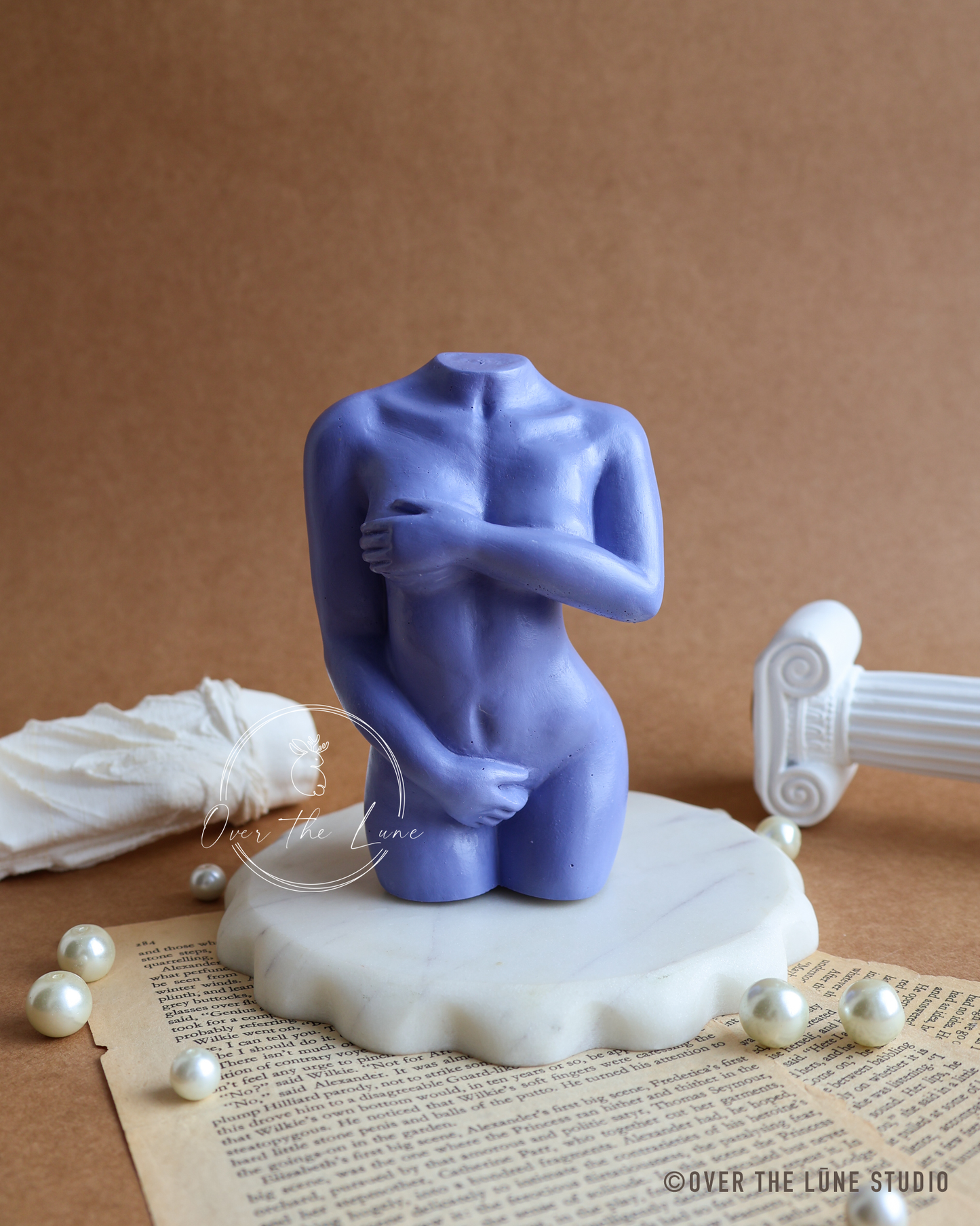A blue-purple abstract female nude sculpture on a white marble base, styled on a vintage book page with scattered pearls, a white draped cloth, and a white classical column fragment against a brown background.