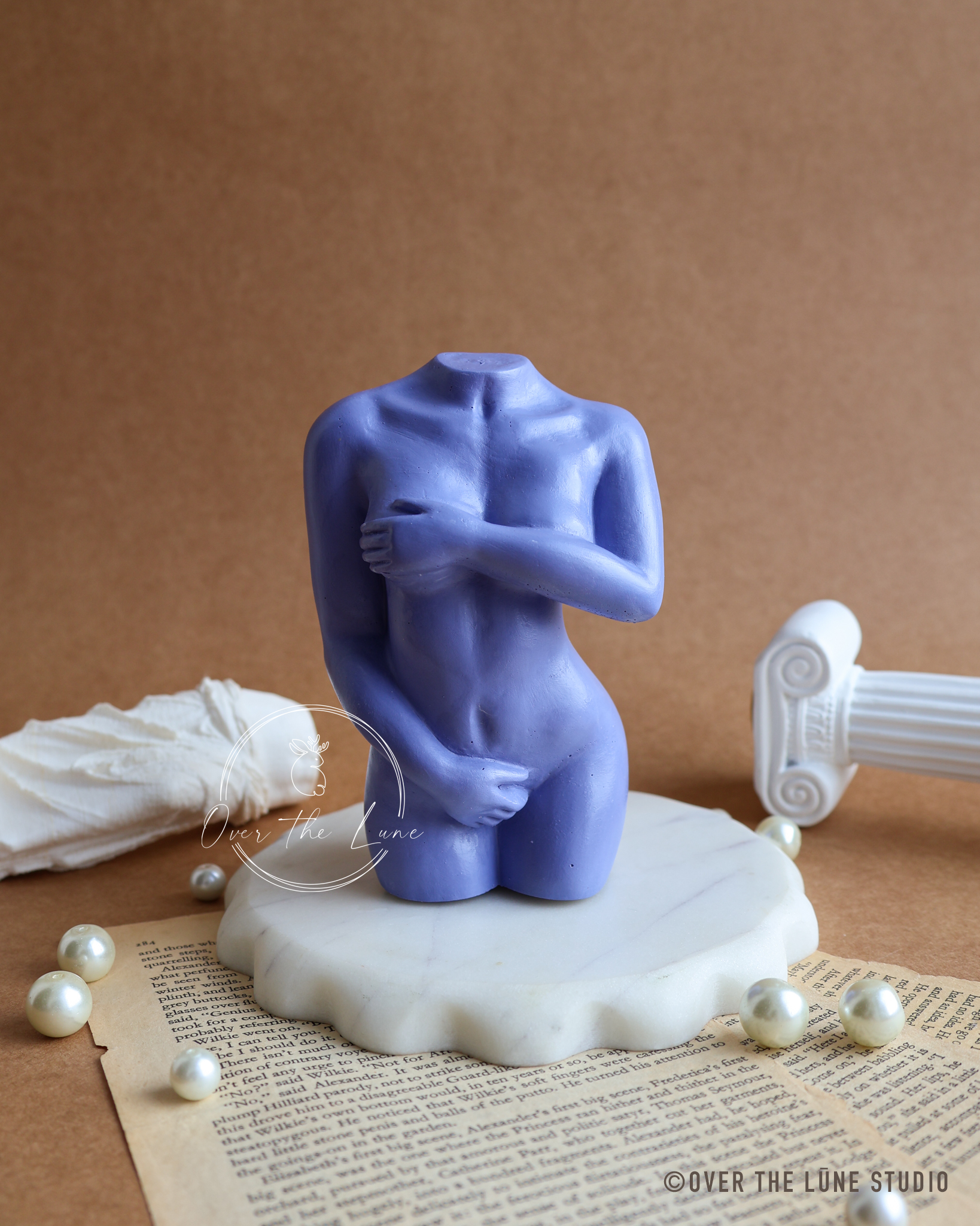 A blue-purple abstract female nude sculpture on a white marble base, styled on a vintage book page with scattered pearls, a white draped cloth, and a white classical column fragment against a brown background.