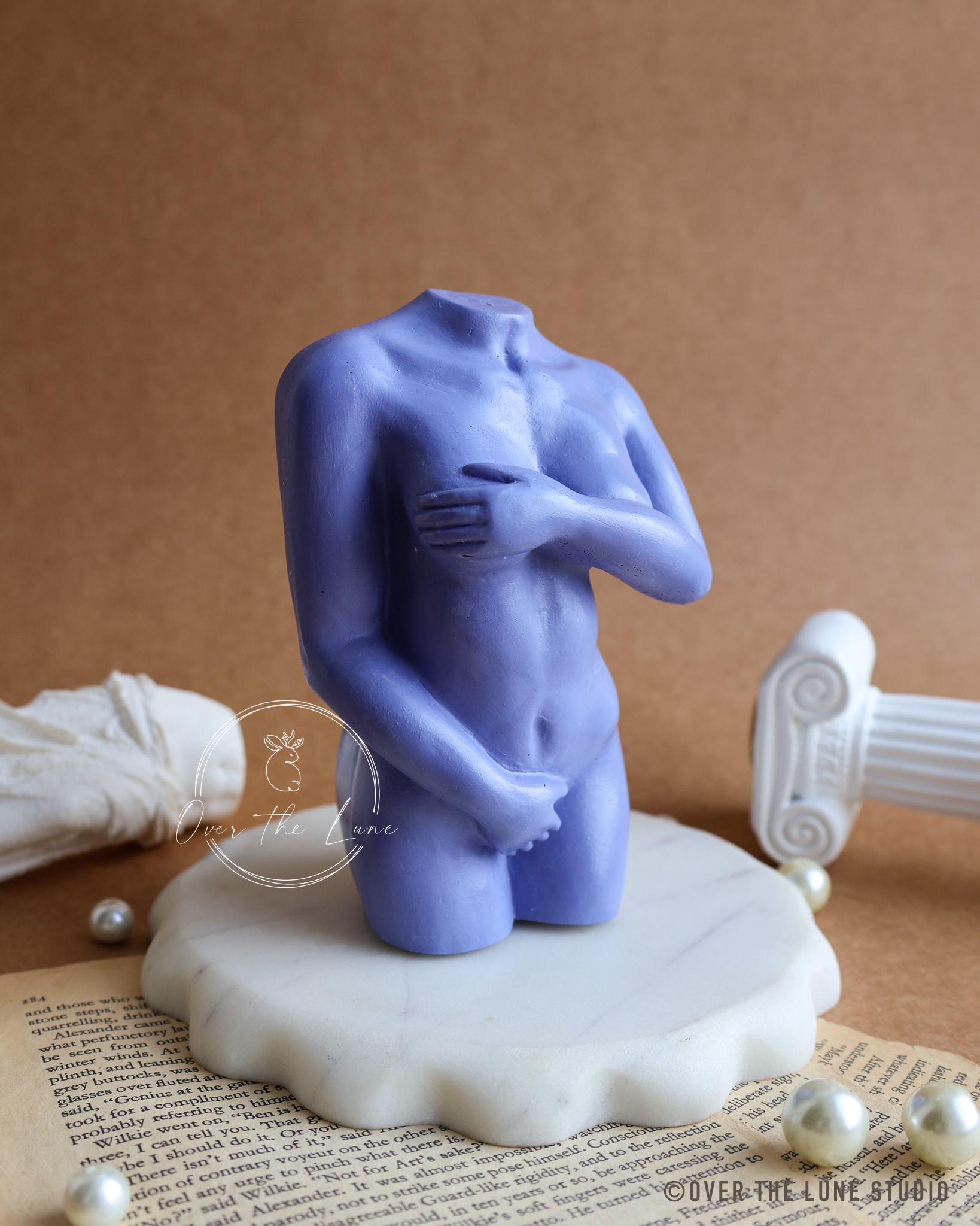 A small lavender-blue headless male nude figurine standing on a white scalloped base, styled on top of an open book with pearl-like beads and a classical column fragment against a brown backdrop.