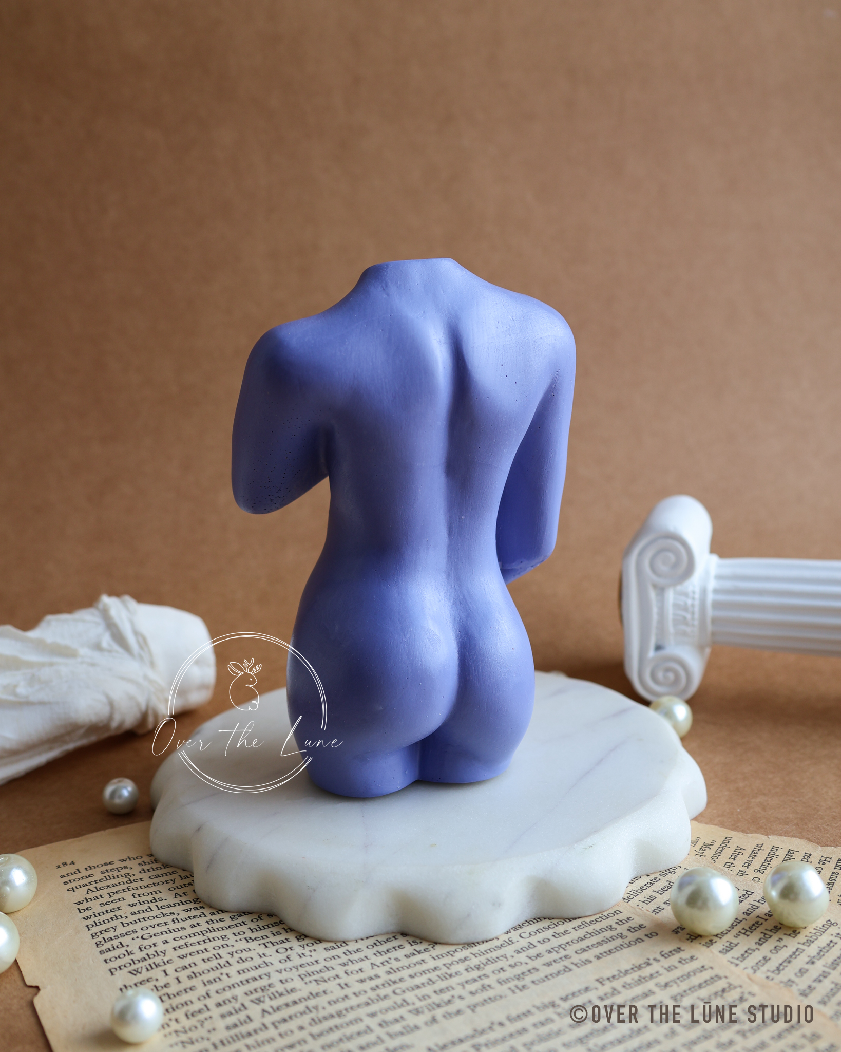 A lavender-blue sculptural female nude torso sculpture displayed from the back on a white marble base, styled with vintage book pages, faux pearls, a draped white cloth, and a classical column fragment against a brown backdrop.