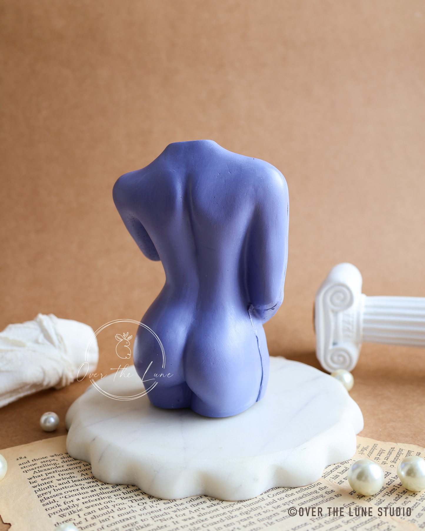 A lavender purple sculptural female torso bust displayed from the back on a white scalloped marble base, set against a warm brown background with decorative props including pearls, an open book, and a classical column fragment.