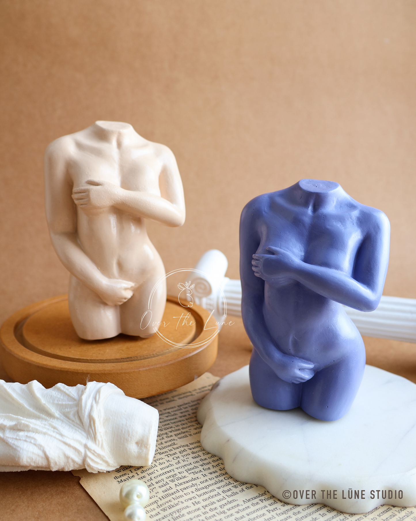 Two modern abstract female nude headless torso sculptures, one in matte beige on a wooden base and the other in matte periwinkle blue on a white stone base, styled on a textured brown background.