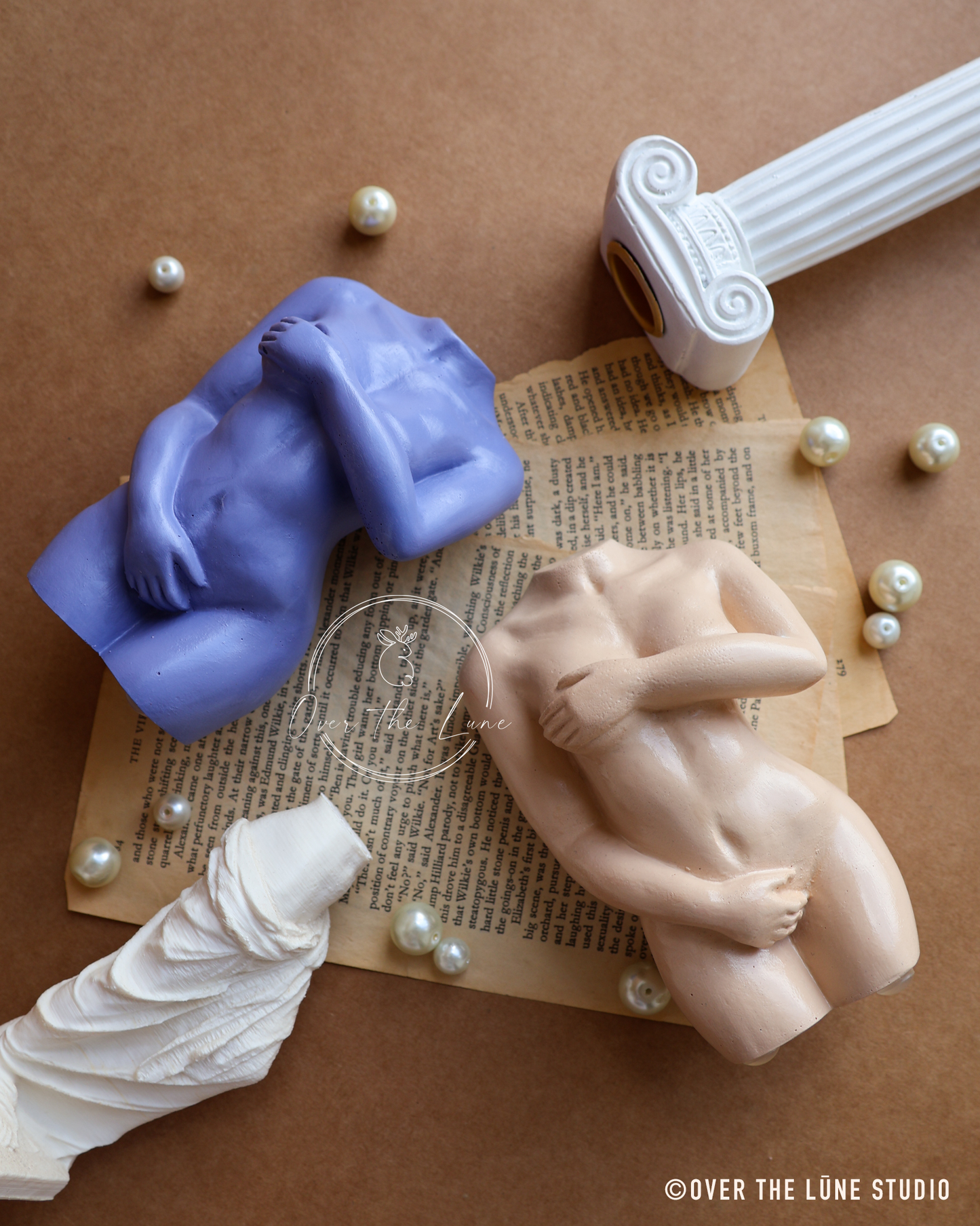 Two matte sculpture-style figurines of nude human forms arranged on vintage book pages with scattered pearls and classical-style decorative props on a brown background. One figurine is lavender-blue and the other is pale peach-beige.