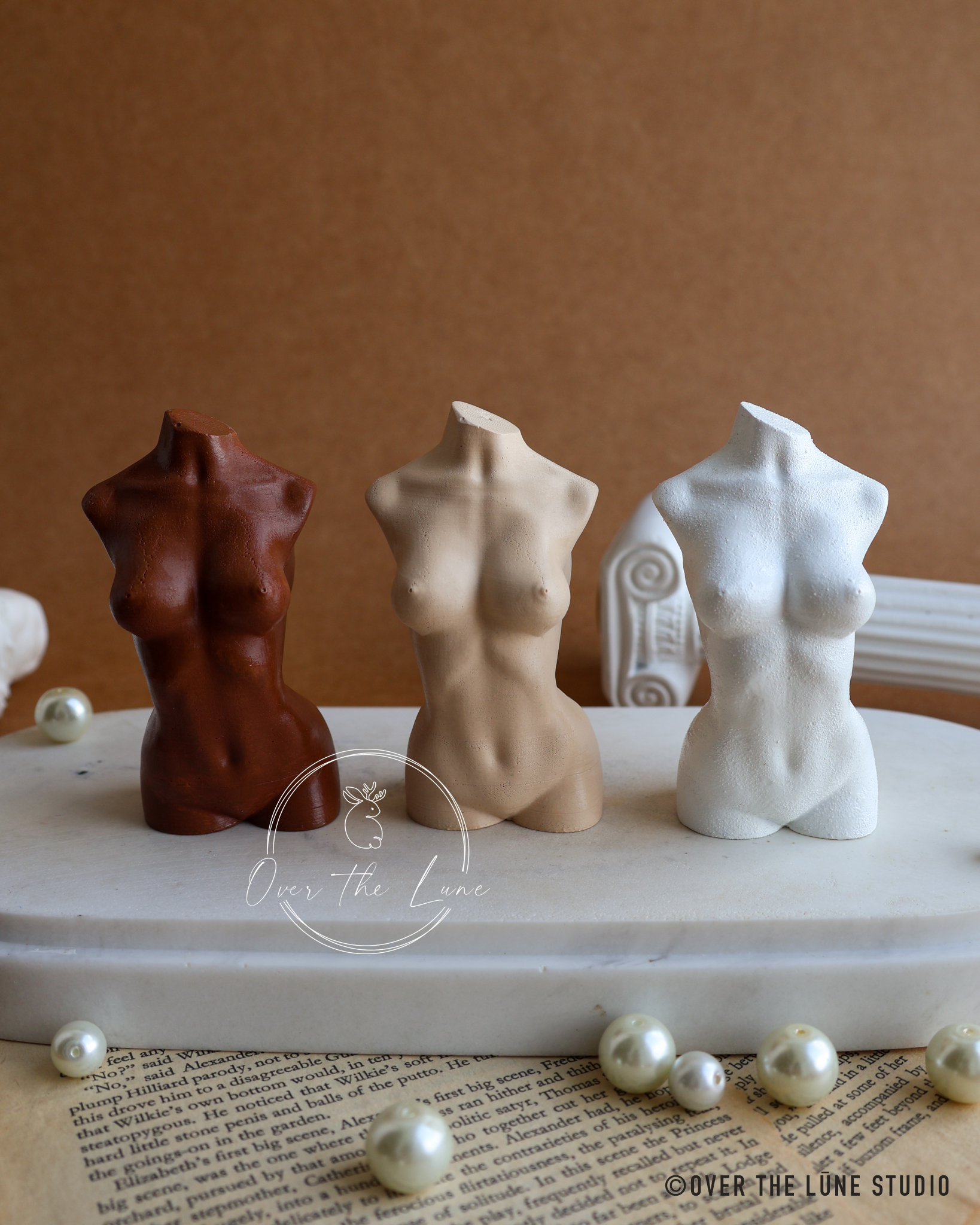Three small female torso figurine in brown, beige, and white are arranged on a white marble slab with pearl-like beads and an open book, against a warm brown background. A watermark reads 'Over The Lune'.