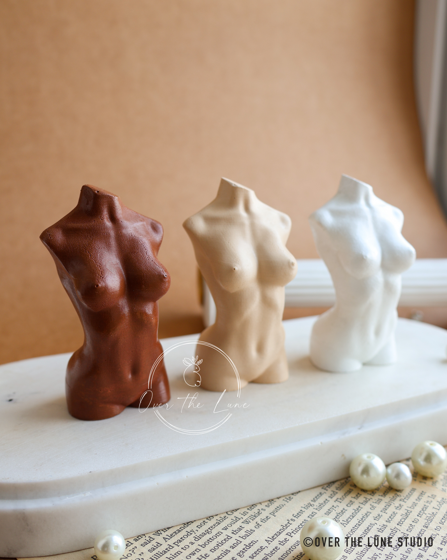 Three small decorative female torso figurines in brown, beige, and white, displayed on a white marble slab with pearl beads and a warm tan background.