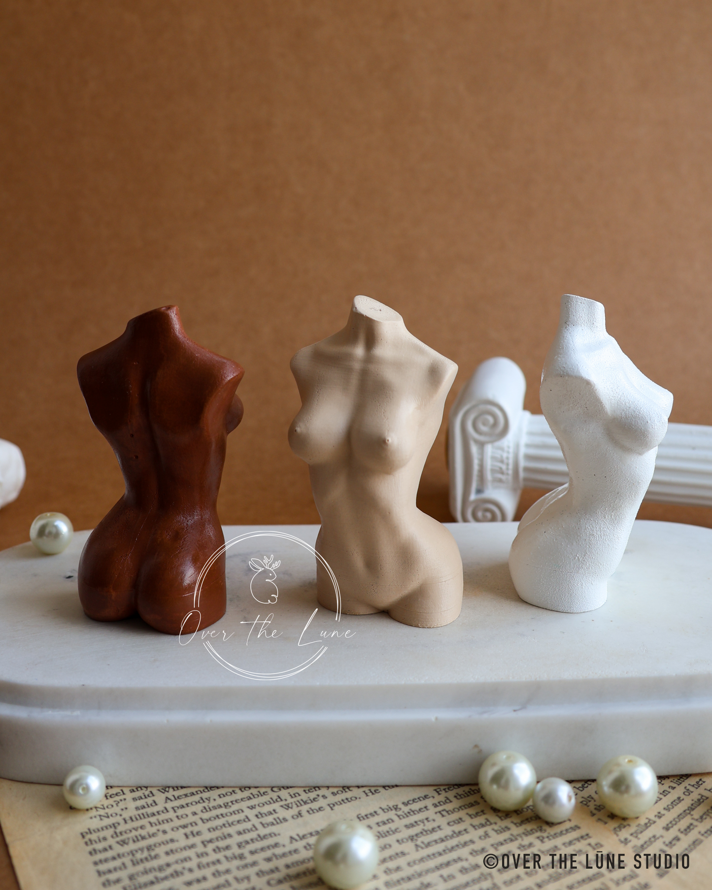 Three small female torso figurines in dark brown, beige, and white ceramic, displayed on a white marble slab with decorative pearls against a warm brown background.