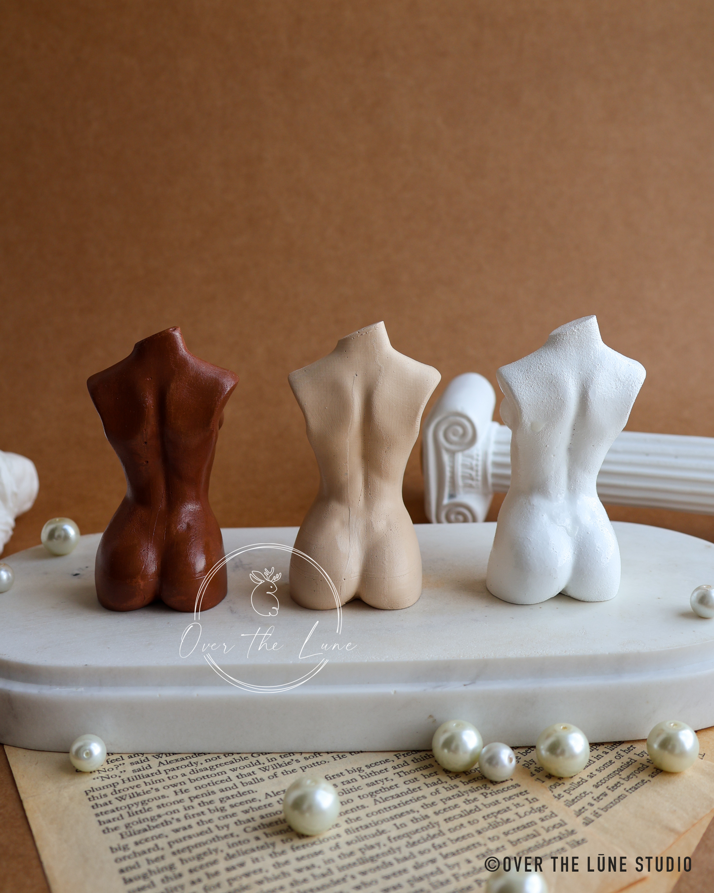 Three miniature torso figurines in brown, beige, and white, displayed on a white pedestal with vintage book pages and pearls against a brown background.