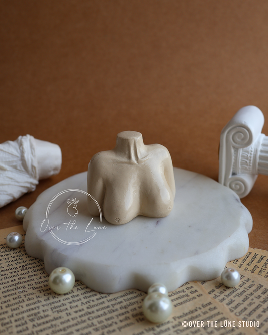 A beige plaster female torso model displayed on a white marble slab, surrounded by faux pearls and classical-style props on a brown background.