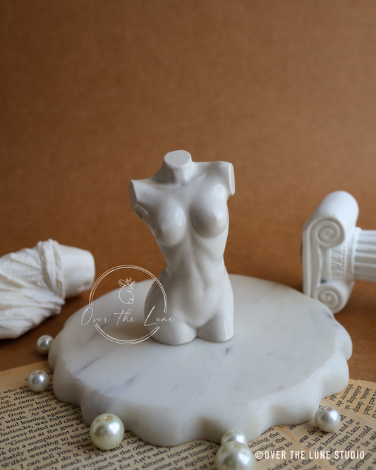 A small white marble female torso mannequin displayed on a scalloped white marble base, with decorative pearls, a rolled fabric piece, and a classical column prop against a warm brown background.