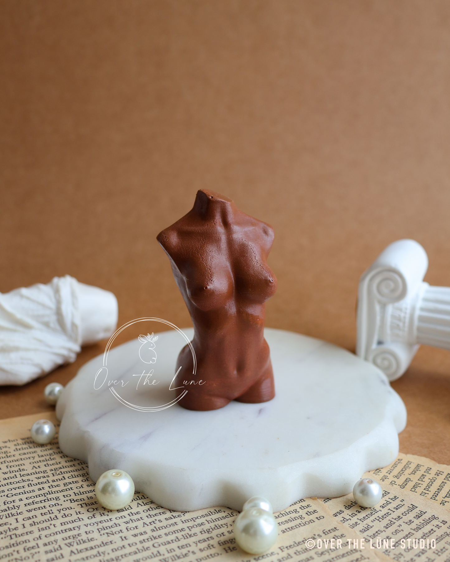 A small brown female torso figurine displayed on a white scalloped marble stand, surrounded by faux pearls and vintage book pages against a warm brown backdrop.