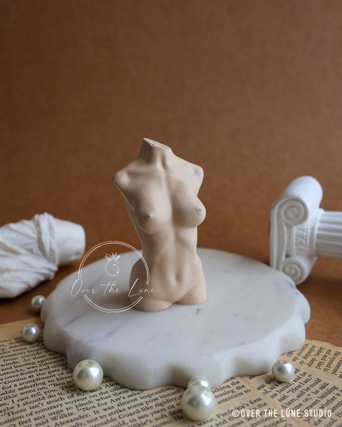 A small beige female torso figurine displayed on a white marble charger plate, surrounded by decorative pearls, a rolled white fabric, and an open book, against a warm brown background.