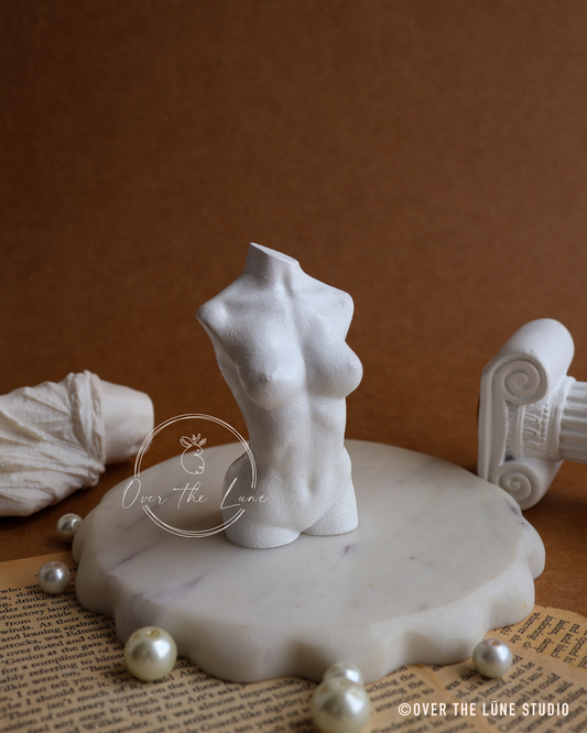 A white female torso figurine displayed on a round white marble pedestal, surrounded by decorative pearls, a draped fabric piece, and a classical column fragment against a warm brown background. A watermark logo for 'Over The Lune' is visible on the image.