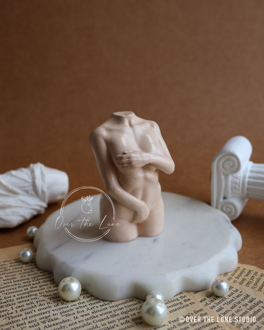A beige ceramic female torso sculpture displayed on a white scalloped marble base, styled with faux pearls, a rolled white fabric, and an open book on a brown background.