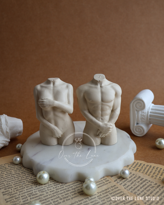 Two beige human torso sculptures on a white marble base, styled on an open book with scattered pearls against a brown background.