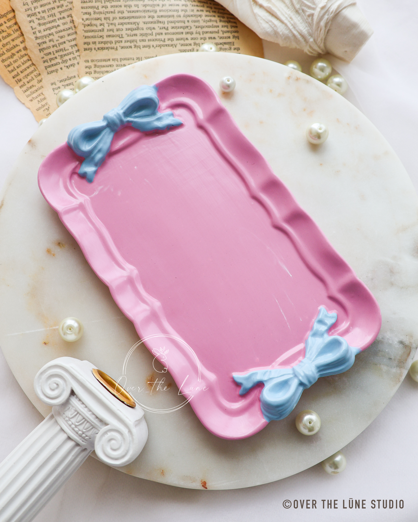 A pink rectangular trinket tray with scalloped edges, decorated with two light blue bow details at the top corners, displayed on a white marble plate with decorative props.