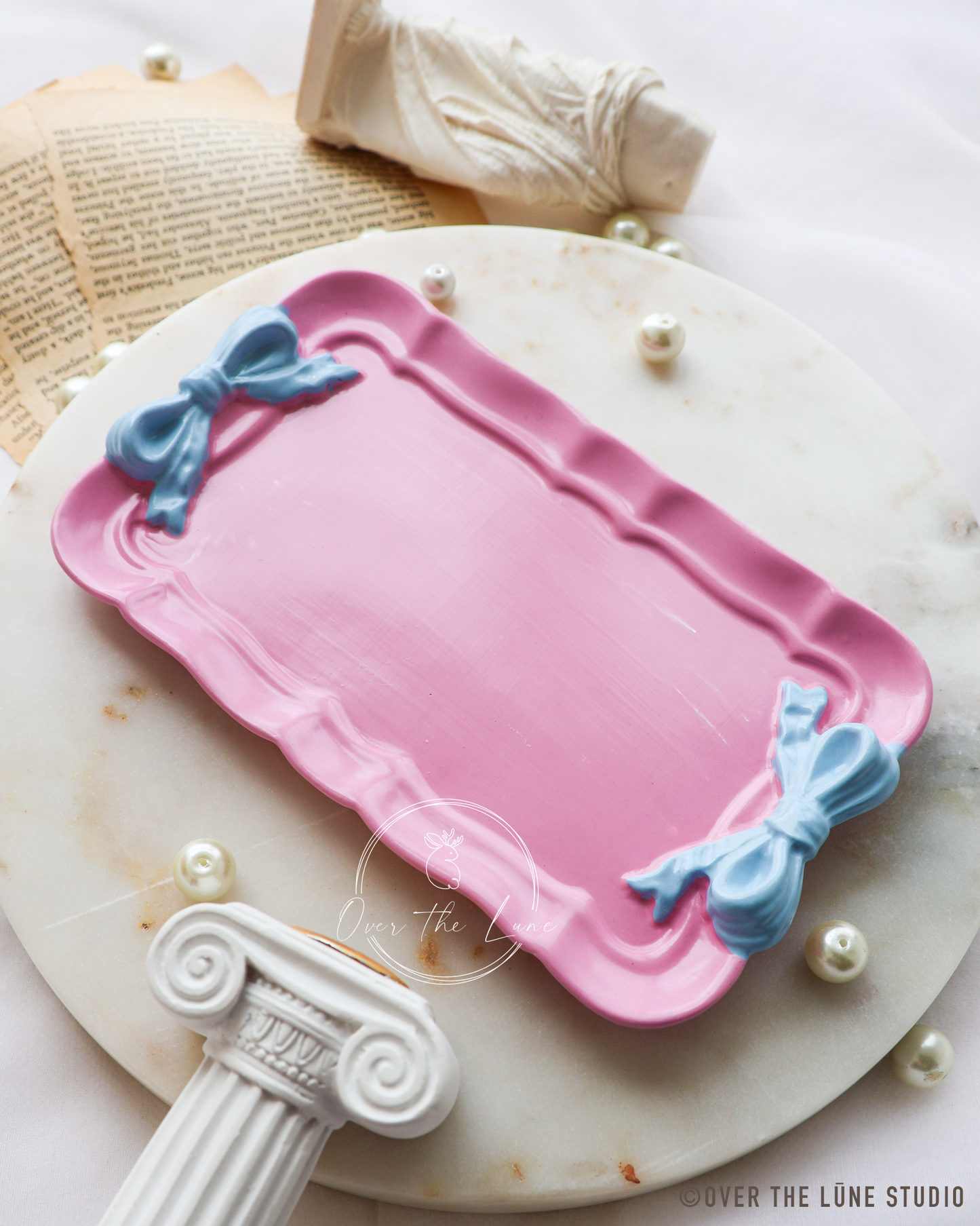 A pink rectangular trinket tray with scalloped edges and two light blue bow decorations on the corners, styled on a round white marble plate with decorative pearls, an open book, and a small white object in the background.