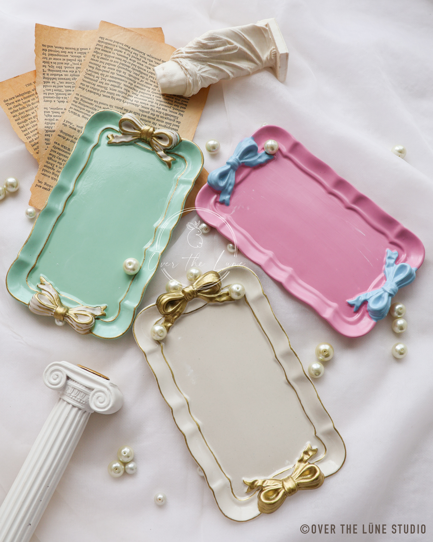 Three decorative rectangular trays in pastel mint green, cream, and pink, each with a scalloped rim, gold bow accents, and pearl details, arranged on a white fabric surface with vintage book pages, pearls, and a small classical column prop.